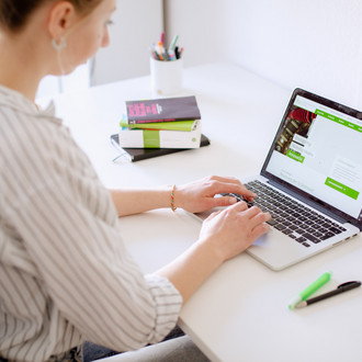 Am Schreibtisch sitzende Studentin tippt auf dem Laptop, daneben liegen Bücher und Stifte.