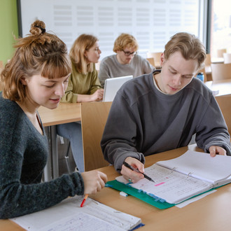 Vier Studierende sitzen im Seminarraum. Zwei sitzen im Vordergrund und arbeiten zusammen.