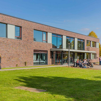 Erich-Brost-Institut: Ein modernes Gebäude mit Flachdach, das rot verklinkert ist und große Fenster und ein großes Vordach hat. Davor eine große Wiese. Menschen sitzen auf einer Bank vor dem Gebäude.