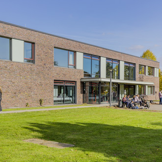 Erich-Brost-Institut: Ein modernes Gebäude mit Flachdach, das rot verklinkert ist und große Fenster und ein großes Vordach hat. Davor eine große Wiese. Menschen sitzen auf einer Bank vor dem Gebäude.