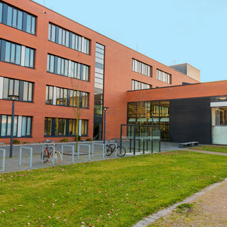 Seminarraumgebäude: Ein rotes dreistöckiges Gebäude mit schwarz gerahmten Fenstern, von der Seite fotografiert. Davor eine Wiese und Fahrradständer sowie Straßenlaternen.