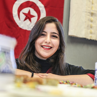 Eine internationale Studierende lächelt in die Kamera. Im Hintergrund hängt eine Tunesienflagge.