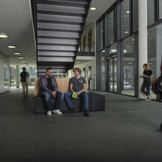 Zwei Studierende sitzen auf einer Bank und unterhalten sich. Andere laufen vorbei.