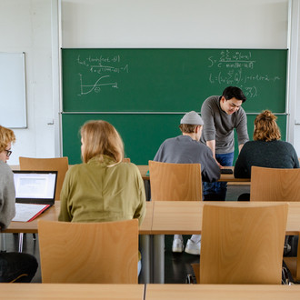 Vier Studierende im Seminarraum sitzen und arbeiten zusammen mit dem Dozierenden im Hintergrund.