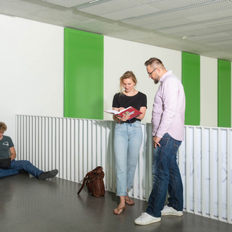 Studierende stehen mit Büchern vor einem Geländer, ein anderer sitzt auf dem Boden.