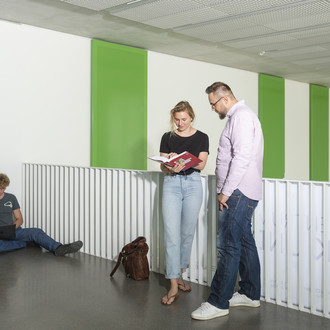 Studierende stehen mit Büchern vor einem Geländer, ein anderer sitzt auf dem Boden.