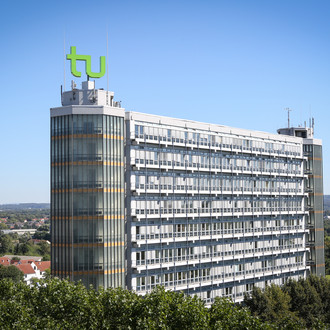 Mathematikgebäude mit Mathetower: Ein hohes Gebäude mit vielen Fenstern und je einem Turm an beiden Seiten. Auf dem linken Turm ein grünes TU-Logo. Im Hintergrund Häuser und Bäume.