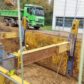 Hinter einem Bauzaun ist ein langer rechteckiger Aushub, in dem ein Bauarbeiter mit gelber Schutzjacke und Helm arbeitet. Gelbe Metallstützen stützen die Seiten des Aushubs ab. Auf der gegenüberliegenden Seite der Baustelle parkt ein dunkelgrüner LKW.