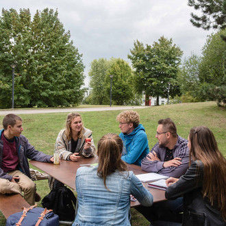 Sechs Studierende sitzen an einer Sitzgruppe auf der Wiese mit Bäumen im Hintergrund.