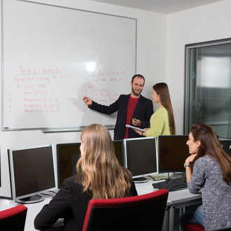 Im Hintergrund stehen zwei Personen vor einem Whiteboard mit Schrift. Die Person links zeigt mit einem Stift auf das Whiteboard. Die Person daneben hört zu und hat einen Block in der Hand. Im Vordergrund sieht man zwei weitere Personen von hinten, die vor PC-Bildschirmen sitzen.