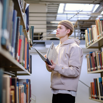 Studierender steht vor Bücherregal in der Bibliothek und sucht nach Büchern.