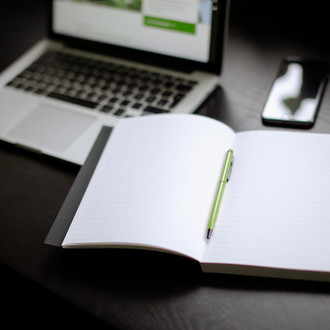 Eingeschalteter Laptop, Smartphone und Notizbuch mit Stift auf einem Tisch liegend.