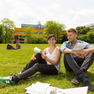 Zwei Studierende sitzen auf einer Wiese, im Hintergrund die Mensa