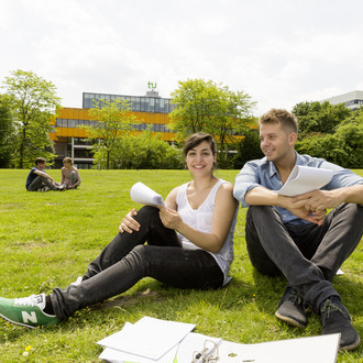 Zwei Studierende sitzen auf einer Wiese, im Hintergrund die Mensa