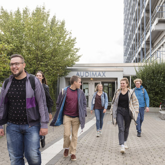 Sechs Studierende laufen teilweise zueinander gewandt aus dem Audimaxgebäude heraus.