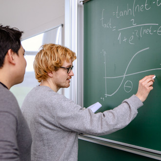 Zwei männliche Studierende an der Tafel, einer schreibt, der andere guckt dahinterstehend zu