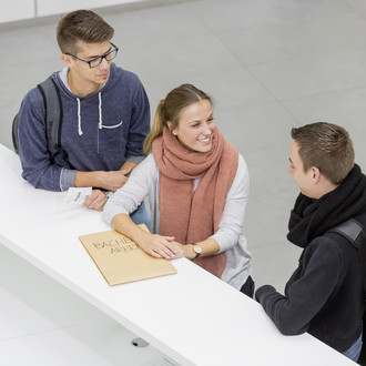 Drei Personen stehen neben einenander an einer Theke. Die Person in der Mitte lacht die Person rechts an und hat ihre Hände auf einen großen Umschlag liegen. Die Person links hat einen kleinen Zettel mit einer Wartenummer in der Hand.