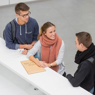 Drei Personen stehen neben einenander an einer Theke. Die Person in der Mitte lacht die Person rechts an und hat ihre Hände auf einen großen Umschlag liegen. Die Person links hat einen kleinen Zettel mit einer Wartenummer in der Hand.