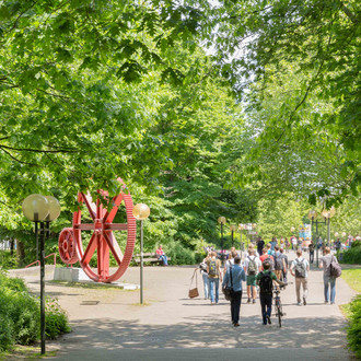 Studierende sind im Sommer auf dem Campus Nord, rote Zahnräder im Hintergrund.