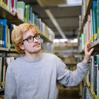 Student steht suchend zwischen Bücherregalen.