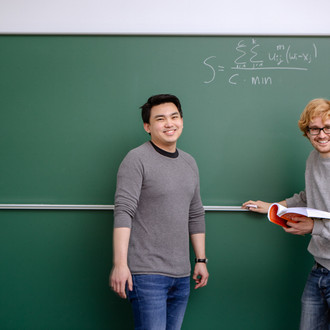 Zwei Studierende stehen vor der Tafel, lächeln in die Kamera