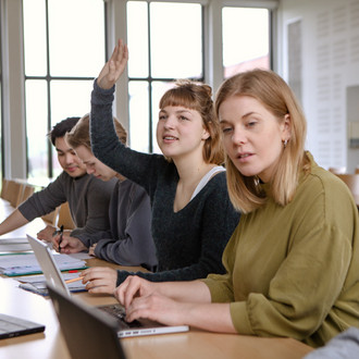 Studierende im Seminarraum sitzen in einer Tischreihe vor ihren Laptops. Eine Studierende meldet sich.