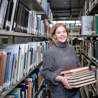 Studierende steht mit einem Stapel Bücher in den Händen zwischen den Bücherregalen der Bibliothek.