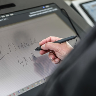 Bildschirm eines Hörsaalpults, an dem jemand mit einem Stift in der Hand schreibt.