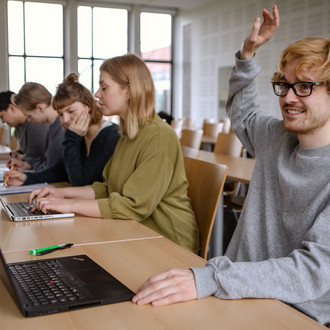 Studierende im Seminarraum sitzen in einer Tischreihe vor ihren Laptops. 