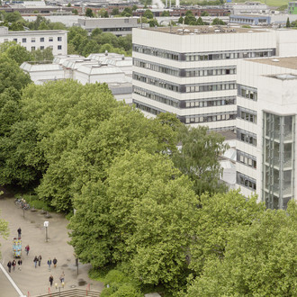 BCI: Rechts im Bild befinden sich zwei hohe graue Gebäude mit vielen Fenstern der Fakultät Bio- und Chemieingenieurwesen. Davor und weiter links im Bild Bäume.