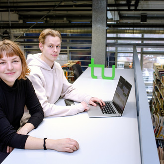 Zwei Studierende sitzen an einem langen Tisch vor einem Laptop in der Bibliothek.