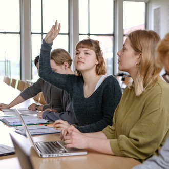 Studierende im Seminarraum sitzen in einer Tischreihe vor ihren Laptops. Eine Studierende meldet sich.