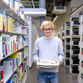 Student trägt Bücher in der Bibliothek und guckt lächelnd in die Kamera.