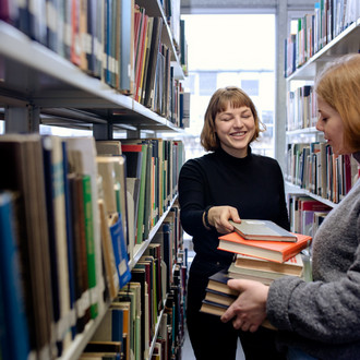 Zwei Studierende stehen zwischen zwei Bücherregalen mit Büchern in der Hand in der Bibliothek.