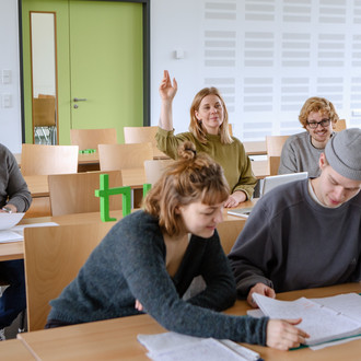 Studierende im Seminarraum sitzen an den Tischen. Eine Person meldet sich. 