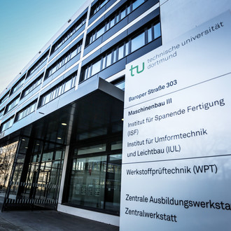 Maschinenbau-Gebäude III: Ein modernes grau-schwarzes Gebäude aus Glas mit Vordach, seitlich von unten fotografiert. Rechts vor dem Gebäude ein Schild mit der Aufschrift "Technische Universität Dortmund, Baroper Straße 303, Maschinenbau 3" und weiteren Instituten und Einrichtungen.