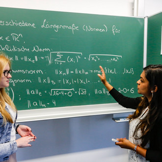 Zwei Personen stehen vor einer Tafel, auf die mit Kreide Formeln geschrieben sind. Die Person rechts zeigt mit dem Arm auf die Tafel und spricht. Die Person links sieht die Person rechts an.