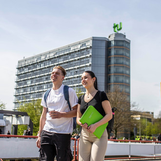 Ein junger Mann und eine junge Frau laufen über die Mensabrücke. Im Hintergrund sieht man den Mathetower mit TU-Logo und das Mensagebäude.