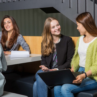 Zwei Studierende unterhalten sich und sitzen gemeinsam auf einer Bank mit einem Laptop.
