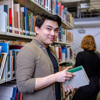 Student steht vor Bücherregal mit einem Buch in der Hand und lächelt in die Kamera.