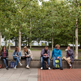 Studierende sitzen auf einer langen Bank auf dem Campus Süd und schauen in Bücher.