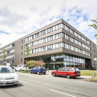 ITMC: Ein großes, braun-rot verklinkertes Gebäude mit Flachdach aus der Froschperspektive fotografiert. Davor eine Straße, auf der ein Auto fährt. Andere Autos stehen am Straßenrand hintereinander geparkt.