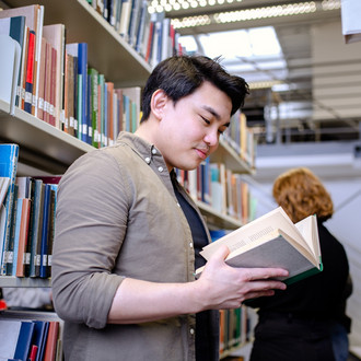 Student steht zwischen zwei Bücherregalen und schaut in ein Buch.