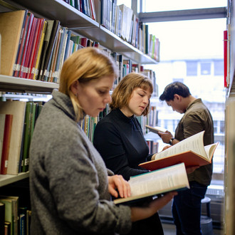 Studierende stehen lesend vor Bücherregalen in der Bibliothek.