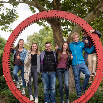 Studierende haben sich in einem Zahnrad aufgestellt und zeigen ihre "Daumen hoch!"
