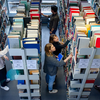 Studierende stehen suchend an mehreren Bücherregalen in der Bibliothek.