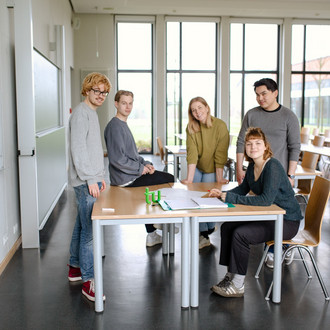Fünf Studierende im Seminarraum stehen um einen Tisch und arbeiten miteinander