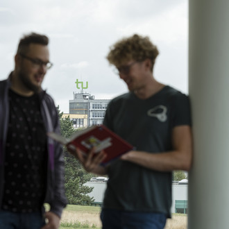 Zwei Studierenden unterhalten sich vor einem großen Fenster, durch das das Logo der TU auf dem Mathetower zu sehen ist.