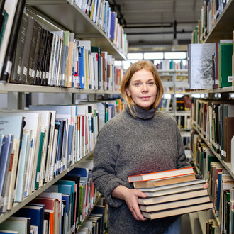 Studentin trägt einen Bücherstapel in den Händen und lächelt in die Kamera.