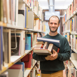 Ein Student steht in der Bibliothek im Gang und hat Bücher auf dem Arm.